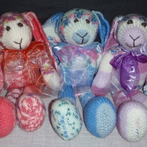 Puede incluir: Tres conejos de punto con orejas largas, cada uno con una cinta de color diferente. Los conejos son rosa, azul y morado con detalles blancos.