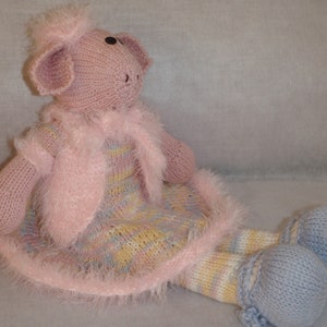 May include: A knitted stuffed animal with a pink head, ears, and arms, wearing a fuzzy pink vest over a multicolored pastel dress. The figure has long legs with striped patterns and light blue rounded feet, featuring black eyes and stitched details.