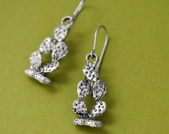 May include: A pair of silver earrings shaped like a cactus plant. The earrings are made of metal and have a textured surface.
