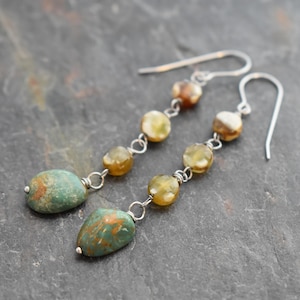 May include: A pair of dangle earrings featuring turquoise-colored, teardrop-shaped stones and small, round, amber-colored beads. The earrings have silver-colored hooks and connecting links. The stones have brown and orange veining.