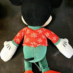 Puede incluir: Un juguete de peluche de Mickey Mouse con un su&eacute;ter rojo con copos de nieve blancos y pantalones verdes. El juguete tiene orejas negras grandes, guantes blancos y zapatos rojos.