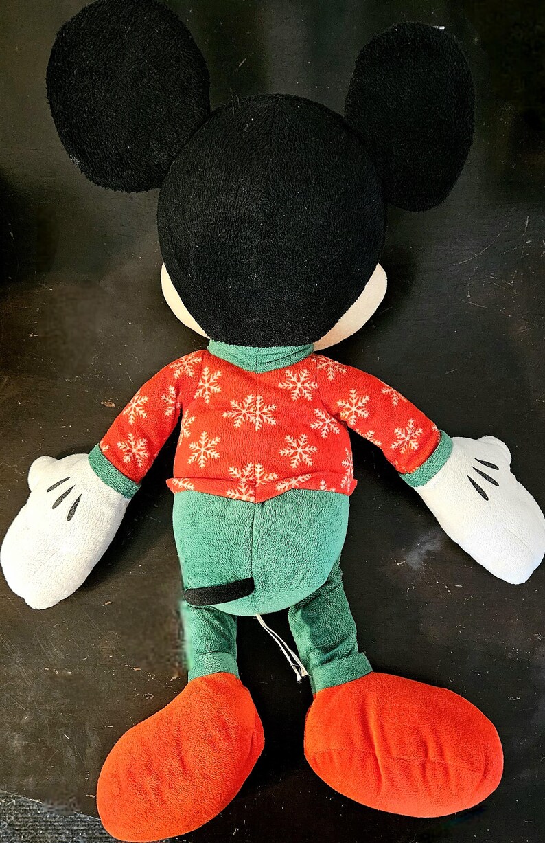 Puede incluir: Un juguete de peluche de Mickey Mouse con un su&eacute;ter rojo con copos de nieve blancos y pantalones verdes. El juguete tiene orejas negras grandes, guantes blancos y zapatos rojos.
