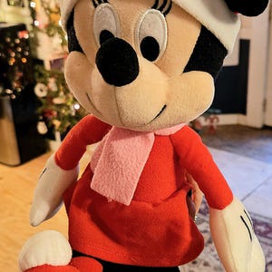 Puede incluir: Un juguete de peluche de Minnie Mouse con un vestido rojo, una bufanda rosa y un lazo rosa. Tiene una cara blanca con ojos negros y una nariz negra.