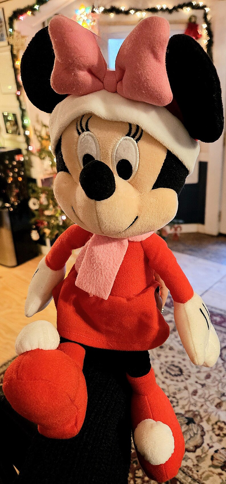 Puede incluir: Un juguete de peluche de Minnie Mouse con un vestido rojo, una bufanda rosa y un lazo rosa. Tiene una cara blanca con ojos negros y una nariz negra.