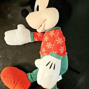 Puede incluir: Un juguete de peluche de Mickey Mouse que lleva un su&eacute;ter rojo y verde con un patr&oacute;n de copo de nieve. El juguete tiene una cara blanca con ojos negros y una gran sonrisa. El juguete lleva zapatos rojos.