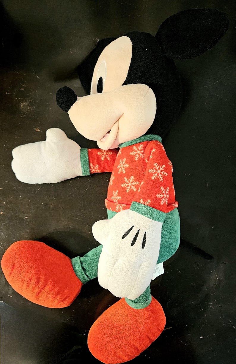 Puede incluir: Un juguete de peluche de Mickey Mouse que lleva un su&eacute;ter rojo y verde con un patr&oacute;n de copo de nieve. El juguete tiene una cara blanca con ojos negros y una gran sonrisa. El juguete lleva zapatos rojos.