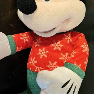 Puede incluir: Un juguete de peluche de Mickey Mouse con un su&eacute;ter navide&ntilde;o rojo y verde con copos de nieve blancos. El juguete tiene una nariz negra y grandes ojos negros.
