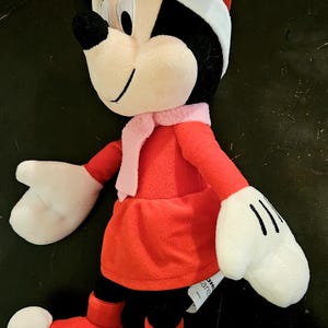 Puede incluir: Un juguete de peluche de Minnie Mouse vestida con un vestido rojo, una bufanda blanca y un gorro de Pap&aacute; Noel rojo con ribete blanco. Lleva botas rojas con pompones blancos. El juguete tiene un lazo rosa en la cabeza.