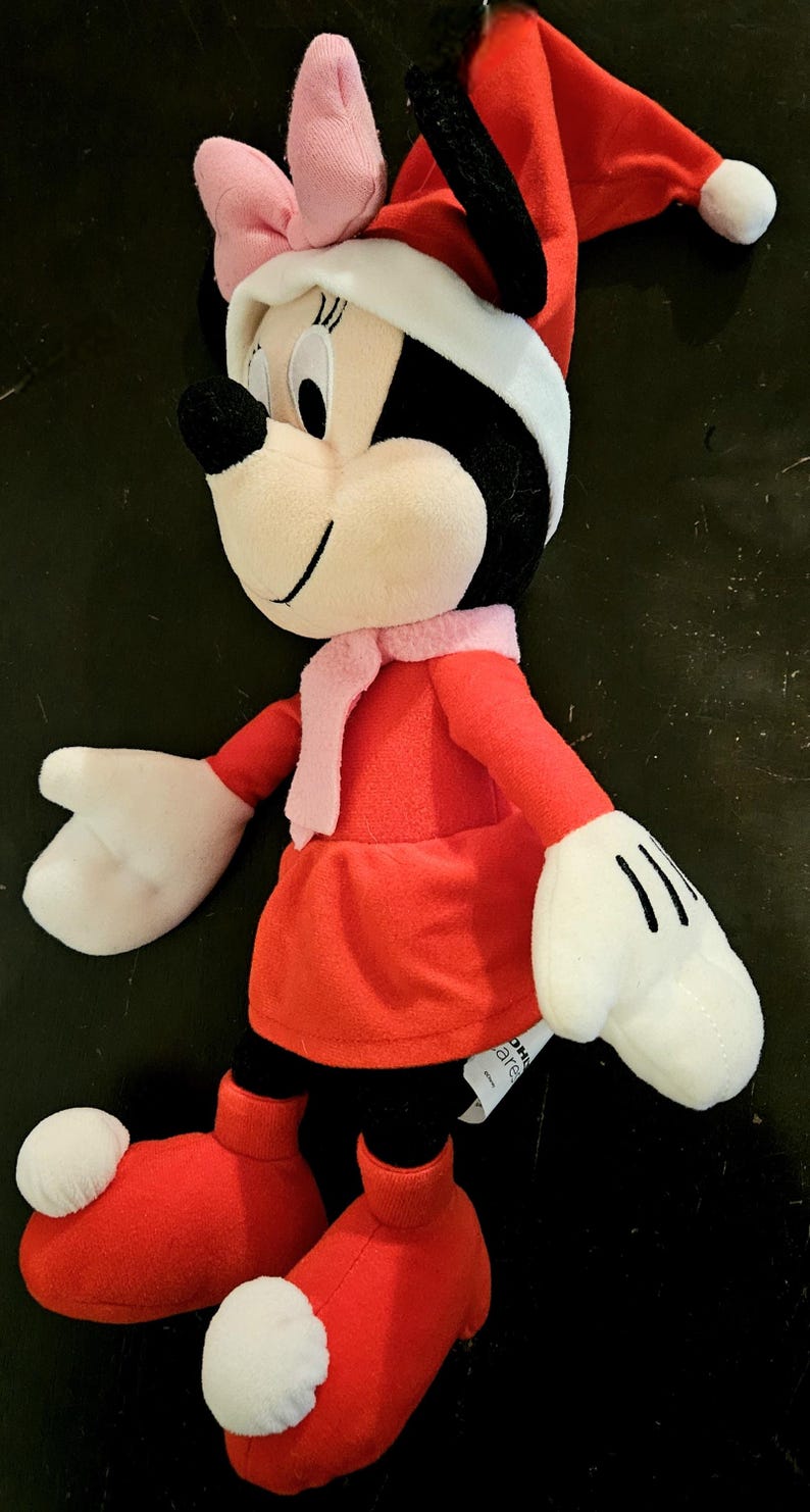 Puede incluir: Un juguete de peluche de Minnie Mouse vestida con un vestido rojo, una bufanda blanca y un gorro de Pap&aacute; Noel rojo con ribete blanco. Lleva botas rojas con pompones blancos. El juguete tiene un lazo rosa en la cabeza.