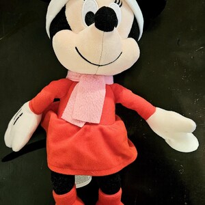 Puede incluir: Un juguete de peluche Minnie Mouse de Navidad rojo y blanco con un lazo rosa y una bufanda rosa.