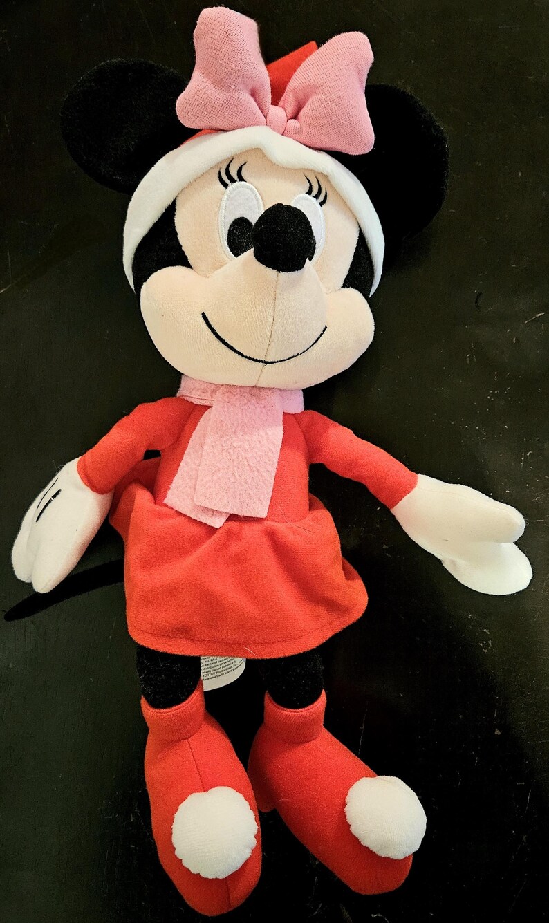 Puede incluir: Un juguete de peluche Minnie Mouse de Navidad rojo y blanco con un lazo rosa y una bufanda rosa.