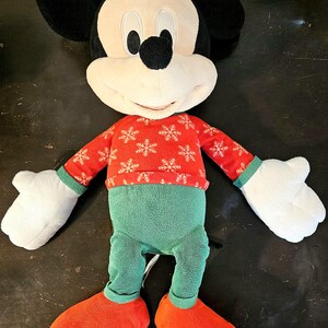 Puede incluir: Un juguete de peluche de Mickey Mouse con un su&eacute;ter rojo y blanco con copos de nieve y pantalones verdes. El juguete tiene una gran sonrisa y est&aacute; de pie sobre una superficie oscura.