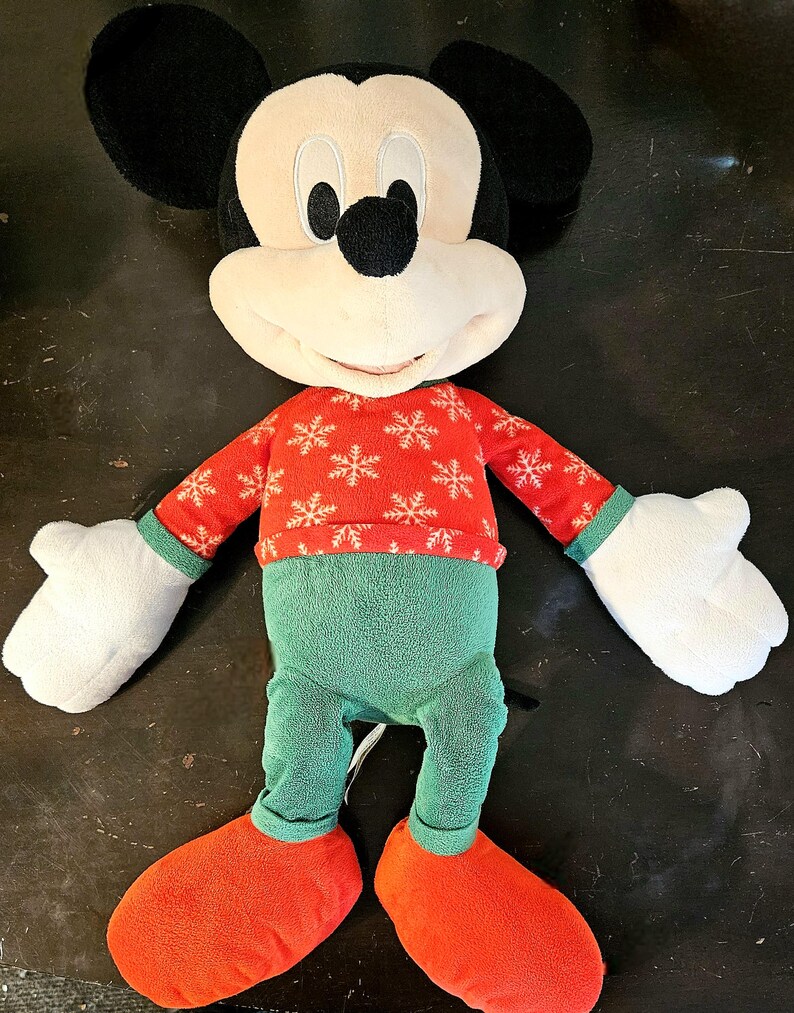 Puede incluir: Un juguete de peluche de Mickey Mouse con un su&eacute;ter rojo y blanco con copos de nieve y pantalones verdes. El juguete tiene una gran sonrisa y est&aacute; de pie sobre una superficie oscura.