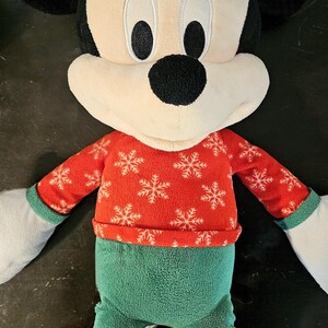 Puede incluir: Un juguete de peluche de Mickey Mouse con un su&eacute;ter rojo y blanco con estampado de copos de nieve y pantalones verdes con zapatos rojos.