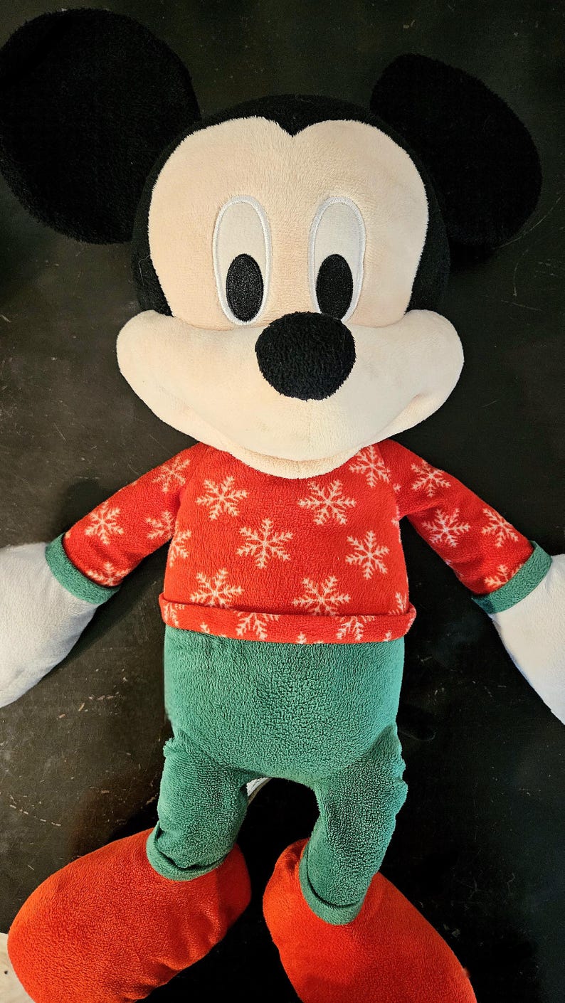 Puede incluir: Un juguete de peluche de Mickey Mouse con un su&eacute;ter rojo y blanco con estampado de copos de nieve y pantalones verdes con zapatos rojos.