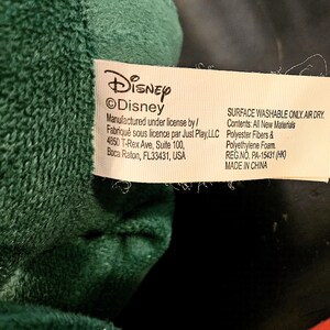 Puede incluir: Una etiqueta blanca con texto negro que dice "Disney &copy;Disney" y "Manufactured under license by/ Fabriqu&eacute; sous licence par Just Play, LLC 4850 T-Rex Ave, Suite 100, Boca Raton, FL33431, USA". La etiqueta tambi&eacute;n dice "SURFACE WASHABLE ONLY. AIR DRY. Contents: All New Materials Polyester Fibers & Polyethylene Foam. REG.NO. PA-15431 (HK) MADE IN CHINA"