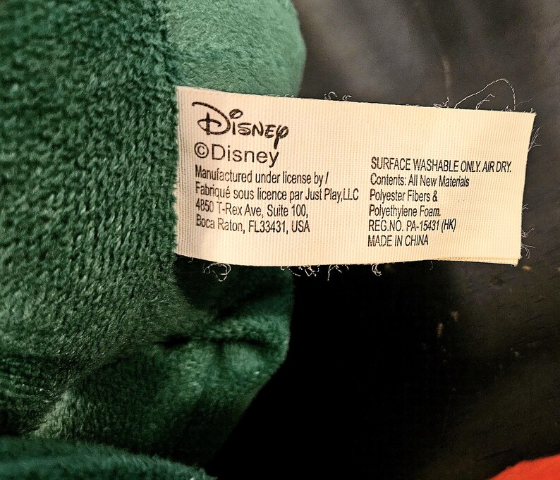Puede incluir: Una etiqueta blanca con texto negro que dice "Disney &copy;Disney" y "Manufactured under license by/ Fabriqu&eacute; sous licence par Just Play, LLC 4850 T-Rex Ave, Suite 100, Boca Raton, FL33431, USA". La etiqueta tambi&eacute;n dice "SURFACE WASHABLE ONLY. AIR DRY. Contents: All New Materials Polyester Fibers & Polyethylene Foam. REG.NO. PA-15431 (HK) MADE IN CHINA"