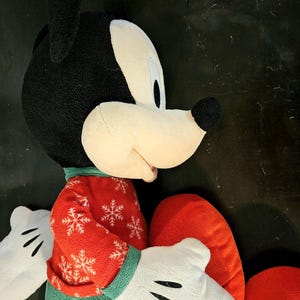 Puede incluir: Un juguete de peluche de Mickey Mouse con un su&eacute;ter navide&ntilde;o rojo y verde. El juguete est&aacute; sentado con las piernas cruzadas y tiene una almohada roja en forma de coraz&oacute;n en su regazo.