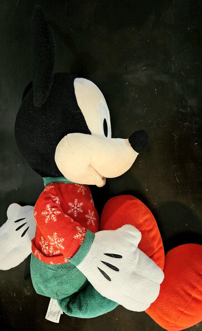Puede incluir: Un juguete de peluche de Mickey Mouse con un su&eacute;ter navide&ntilde;o rojo y verde. El juguete est&aacute; sentado con las piernas cruzadas y tiene una almohada roja en forma de coraz&oacute;n en su regazo.
