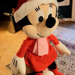 Puede incluir: Un juguete de peluche de Minnie Mouse vestida con un vestido rojo con ribete de piel blanca, un lazo rosa y una bufanda rosa. Lleva botas rojas con ribete de piel blanca.