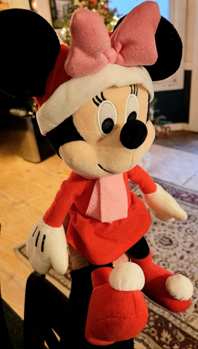 Puede incluir: Un juguete de peluche de Minnie Mouse vestida con un vestido rojo con ribete de piel blanca, un lazo rosa y una bufanda rosa. Lleva botas rojas con ribete de piel blanca.