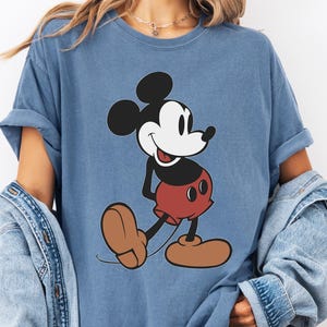 May include: A blue t-shirt featuring a classic cartoon character, Mickey Mouse. The graphic shows Mickey in his iconic pose, with black ears, a white face, red shorts, and brown shoes. The shirt has rolled-up sleeves.
