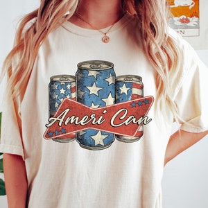 May include: A white t-shirt with a vintage-style graphic of three red, white, and blue American flag-themed cans with the text "Ameri Can" in red and white.