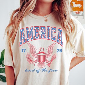 May include: A cream colored t-shirt with a vintage-style graphic of an eagle with the words "AMERICA" and "land of the free" printed on it.