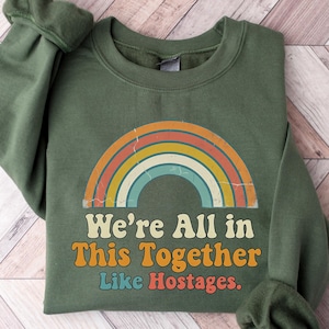 May include: An olive green sweatshirt with a retro rainbow design. The text "We're All in This Together Like Hostages" is printed below the rainbow. The sweatshirt is folded on a light wood-grain surface.