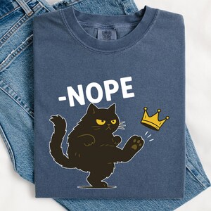 May include: A navy blue t-shirt with the word "-NOPE" in white, and a cartoon black cat kicking a gold crown. The shirt is laid on top of a pair of blue jeans.