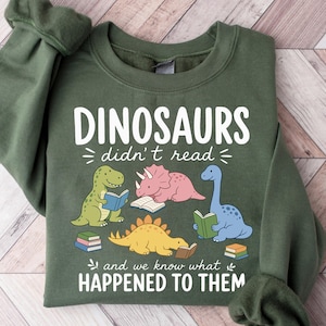May include: An olive green crewneck sweatshirt featuring a graphic of four colorful dinosaurs reading books. The text reads: DINOSAURS didn't read and we know what HAPPENED TO THEM. The design includes a green T-Rex, pink Triceratops, yellow Stegosaurus, and blue long-neck dinosaur.