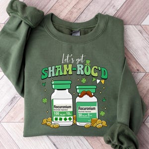 May include: Green sweatshirt with the phrase "Let's get SHAM-ROC'D" in green and white lettering. The design includes two cartoon vials with mustaches and shamrocks, along with gold coins. The sweatshirt is a solid green color.