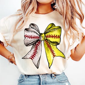 May include: A white t-shirt with a large graphic of a yellow and grey bow made of baseballs. The bow has red stitching.