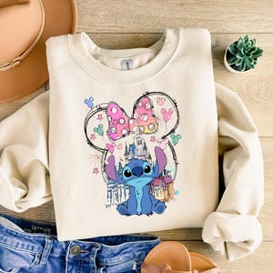 May include: A cream-coloured sweatshirt with a colourful graphic of Stitch from Lilo & Stitch, with a Minnie Mouse bow and a castle. The sweatshirt is paired with blue denim shorts, a tan hat, and sandals.