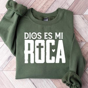 May include: An olive green sweatshirt with the text "DIOS ES MI ROCA" in white block letters. The word "ROCA" features a cross and a heart symbol. The sweatshirt is folded on a light brown and white wooden surface.