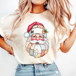 May include: Cream-colored t-shirt featuring a colorful Santa Claus graphic. Santa is depicted with a patchwork beard and hat, adorned with polka dots and holly. The design is festive and evokes a holiday theme.
