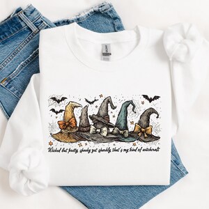 May include: White sweatshirt featuring a graphic of five witch hats in various colors, adorned with bows and surrounded by bats and spiderwebs. The text "Wicked but pretty, spooky yet sparkly, that's my kind of witchcraft" is printed below the hats.