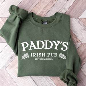 May include: A dark green sweatshirt with the text "PADDY'S IRISH PUB" in white, along with the American flag and the words "SOUTH PHILADELPHIA". The sweatshirt has long sleeves with ribbed cuffs.