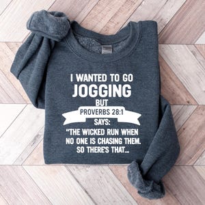 May include: A dark gray sweatshirt with the text "I WANTED TO GO JOGGING BUT PROVERBS 28:1 SAYS: 'THE WICKED RUN WHEN NO ONE IS CHASING THEM. SO THERE'S THAT...'". The sweatshirt is folded on a light brown wooden surface.