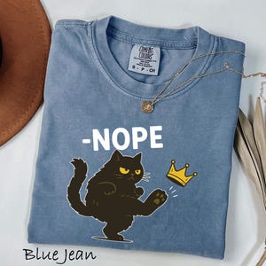 May include: Blue jean colored t-shirt with the word "-NOPE" in white. A black cat with yellow eyes kicks a gold crown. A gold necklace with a pendant is on the shirt. The words "Blue Jean" are at the bottom.