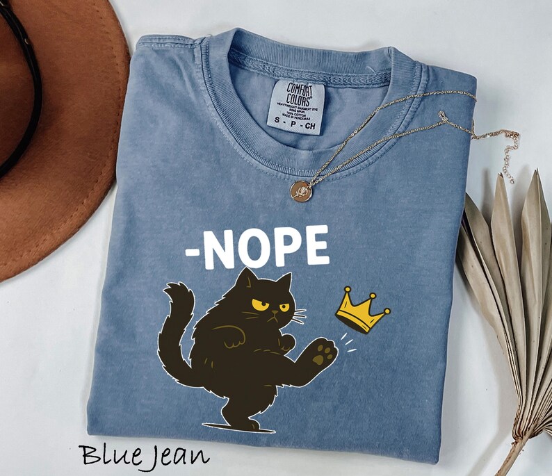 May include: Blue jean colored t-shirt with the word "-NOPE" in white. A black cat with yellow eyes kicks a gold crown. A gold necklace with a pendant is on the shirt. The words "Blue Jean" are at the bottom.