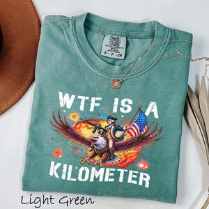 K&ouml;nnte beinhalten: Hellgr&uuml;nes T-Shirt mit dem Text "WTF IS A KILOMETER" und einer Grafik eines Adlers, der einen Soldaten mit Gewehr und amerikanischer Flagge tr&auml;gt. Das Shirt hat einen Rundhalsausschnitt und kurze &Auml;rmel.