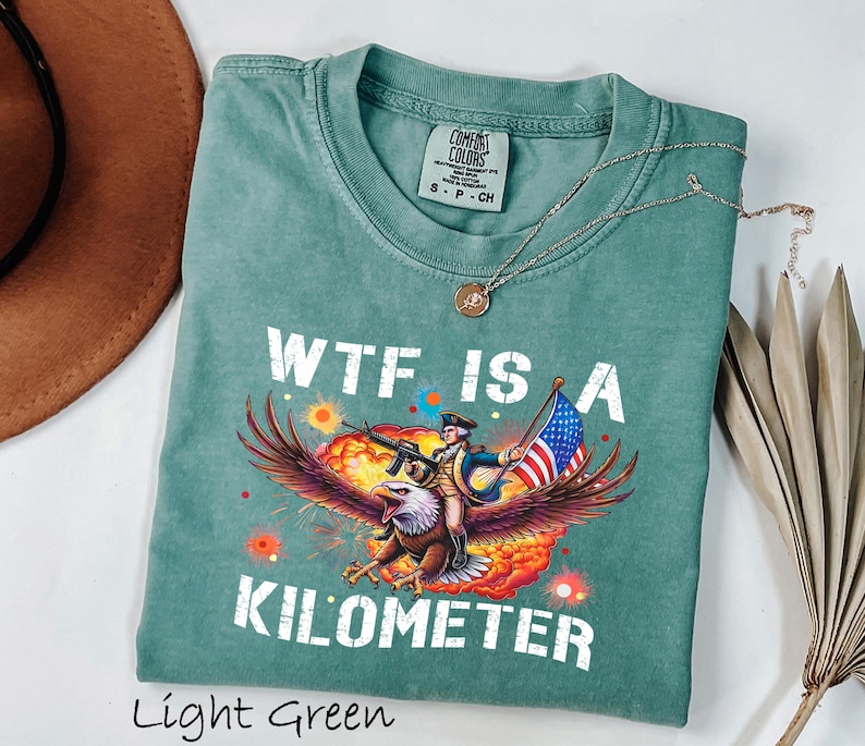 K&ouml;nnte beinhalten: Hellgr&uuml;nes T-Shirt mit dem Text "WTF IS A KILOMETER" und einer Grafik eines Adlers, der einen Soldaten mit Gewehr und amerikanischer Flagge tr&auml;gt. Das Shirt hat einen Rundhalsausschnitt und kurze &Auml;rmel.