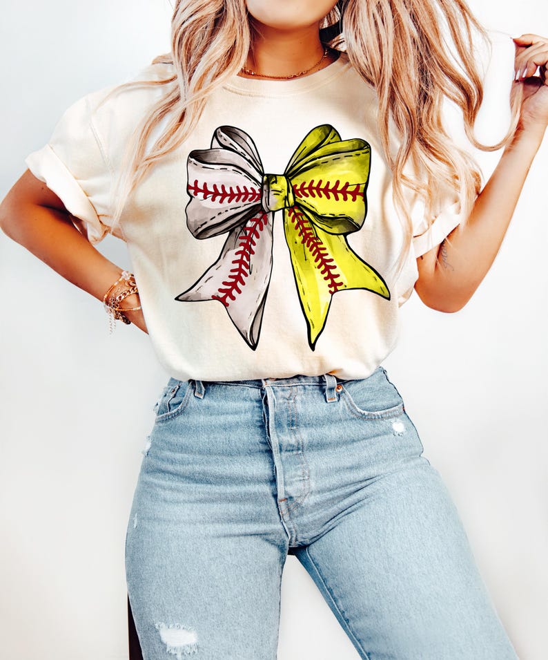 May include: A white t-shirt with a large graphic of a yellow and grey bow made of baseballs. The bow has red stitching.