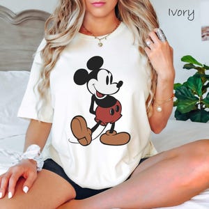 May include: Ivory-colored t-shirt featuring a classic Mickey Mouse graphic. The cartoon character is depicted in black, white, red, and brown. The shirt is a casual, short-sleeved style.