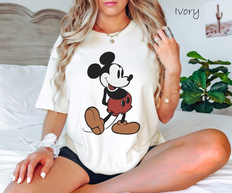 May include: Ivory-colored t-shirt featuring a classic Mickey Mouse graphic. The cartoon character is depicted in black, white, red, and brown. The shirt is a casual, short-sleeved style.
