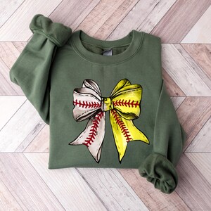 May include: A green sweatshirt with a baseball-themed bow design. The bow is made of two baseballs, one white and one yellow, tied together with red stitching.