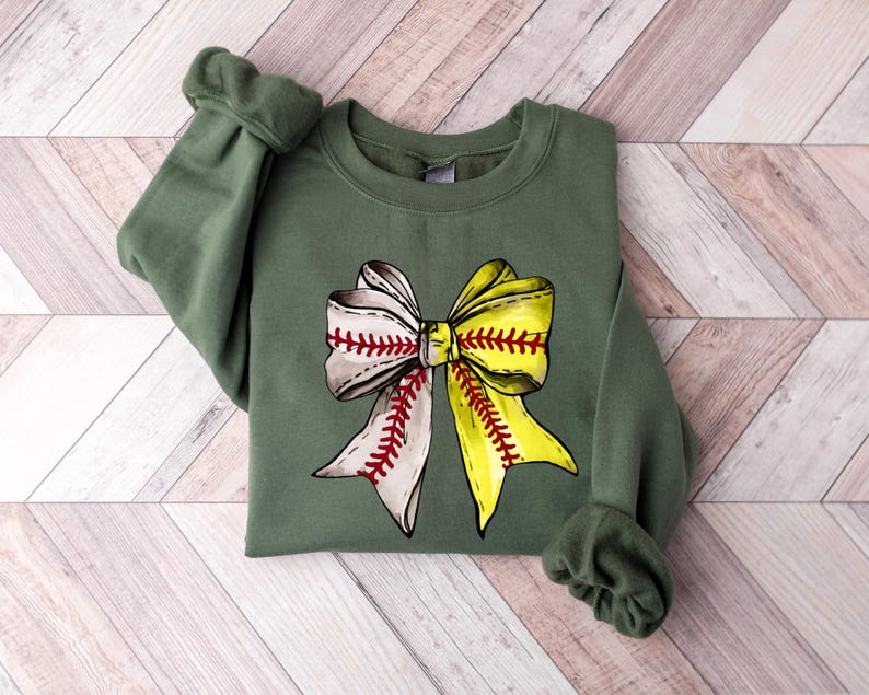 May include: A green sweatshirt with a baseball-themed bow design. The bow is made of two baseballs, one white and one yellow, tied together with red stitching.