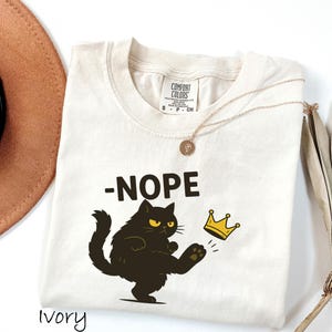 May include: Ivory-colored t-shirt with the word "-NOPE" above a black cat kicking a gold crown. The cat has yellow eyes. A gold necklace and a brown hat are also in the image.