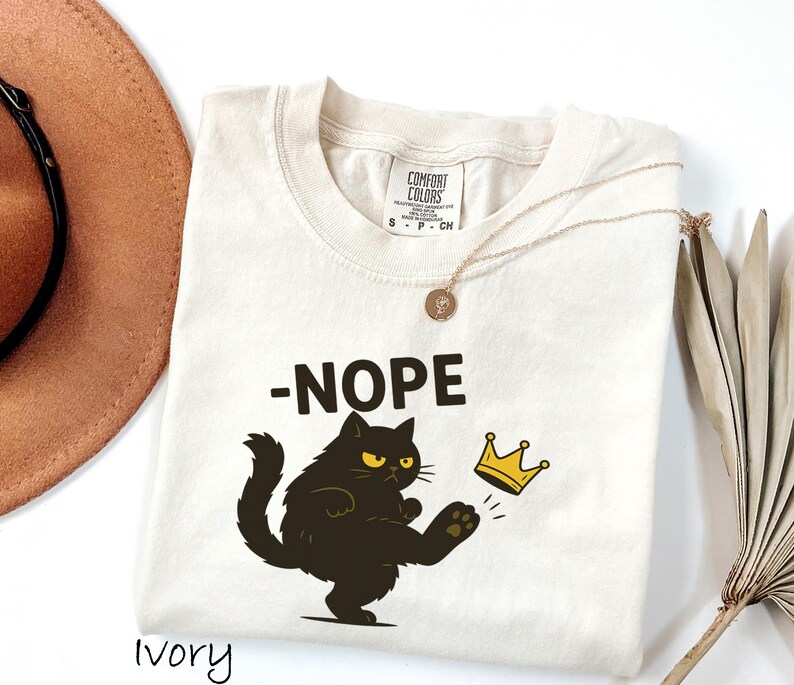 May include: Ivory-colored t-shirt with the word "-NOPE" above a black cat kicking a gold crown. The cat has yellow eyes. A gold necklace and a brown hat are also in the image.